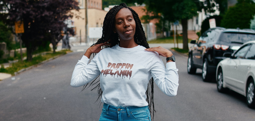 Black femme presenting person standing in street, wearing long sleeve shirt that reads "Melanin Drippin"